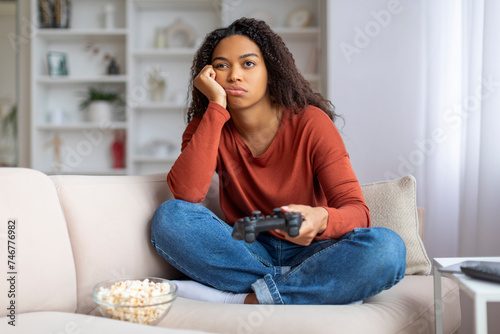 Bored black woman sitting on couch with joystick and looking away