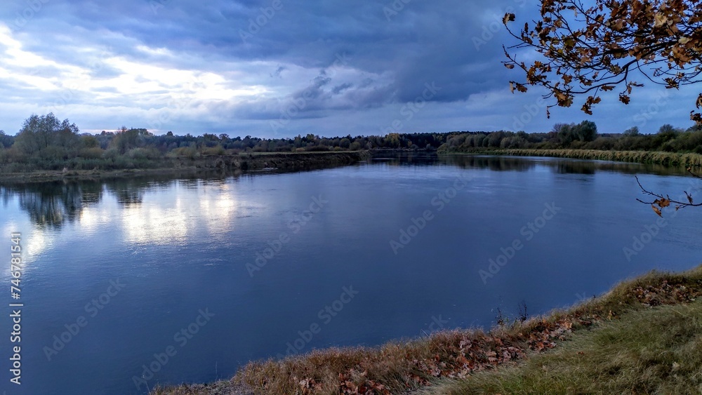 A mixed forest grows on the grassy banks of the river. The river is making a turn. In the fall, the leaves on the trees turn yellow. There are ripples on the water and the reflection of the sunset