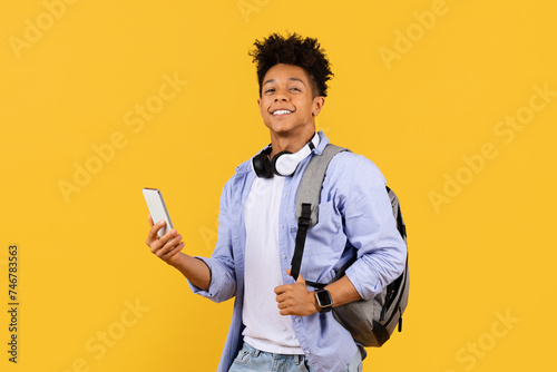 Joyful black male student with headphones and phone on yellow background photo