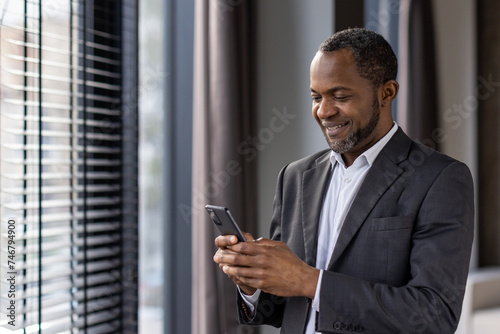 Smiling african american businessman in suit using smartphone by window