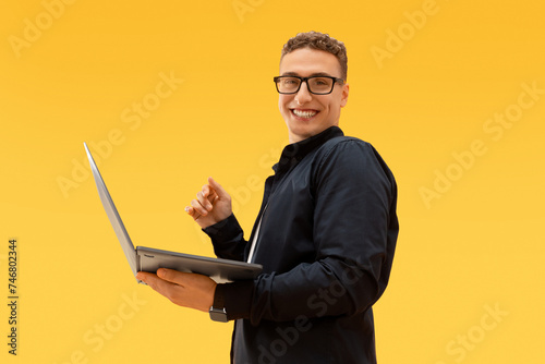 Young guy wearing eyeglasses holding laptop on yellow background