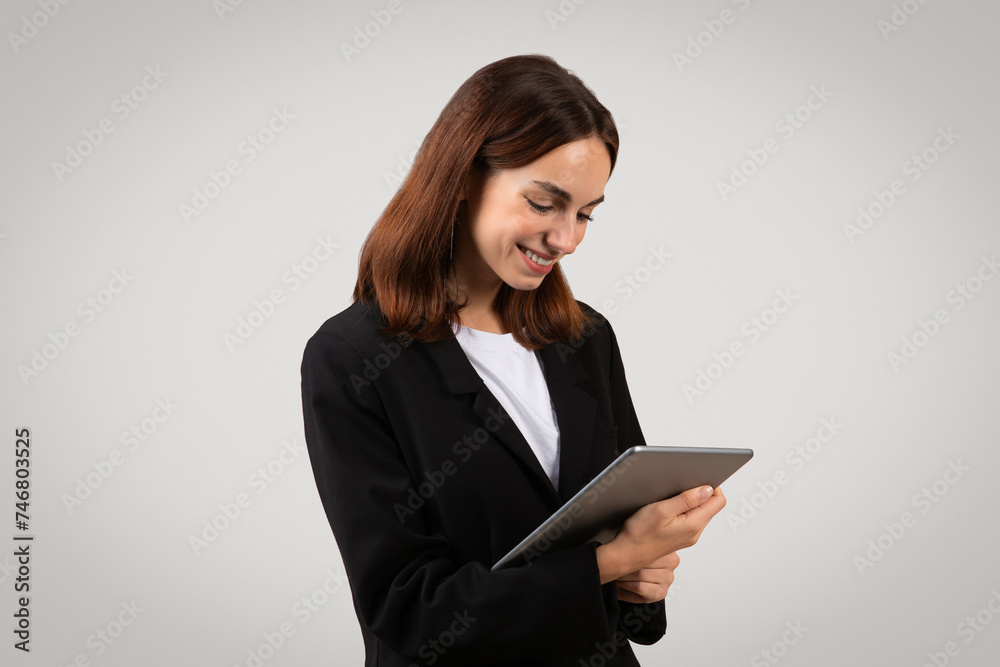 Engaged and attentive young businesswoman in a sleek black suit smiles while focusing on a digital tablet Engaged and attentive young businesswoman in a sleek black suit smiles while focusing on a digital tablet