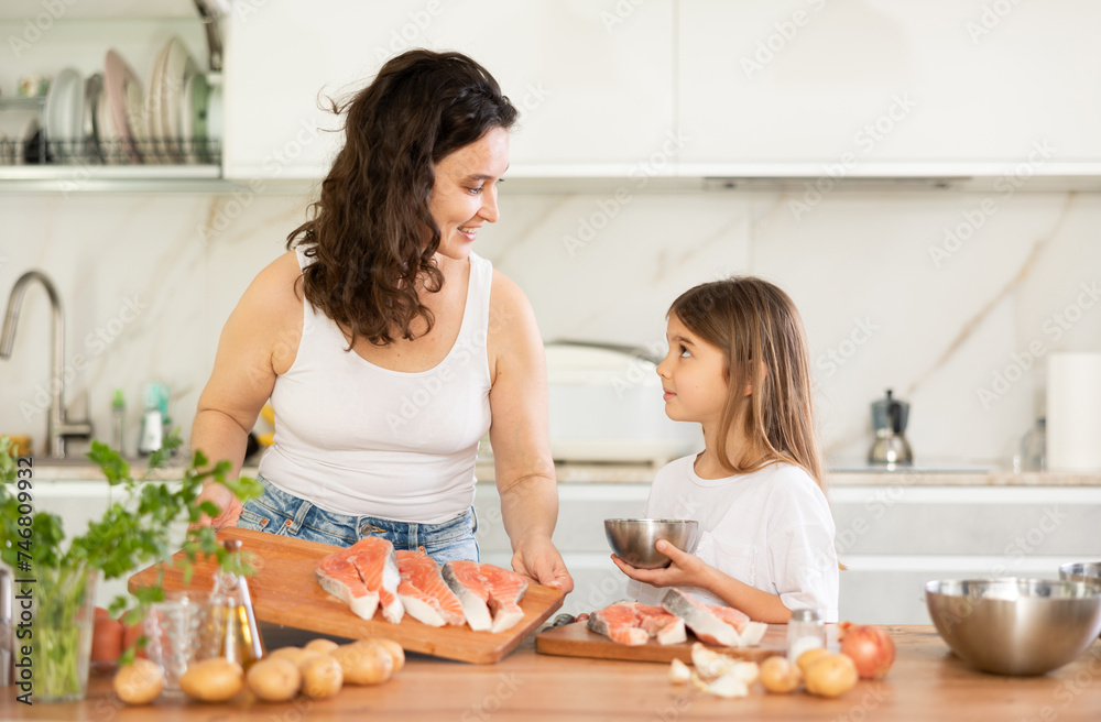 Mom and daughter are preparing festive salmon dish. Family in kitchen practicing cooking salmon dishes. Woman tells girl sequence of actions during cooking