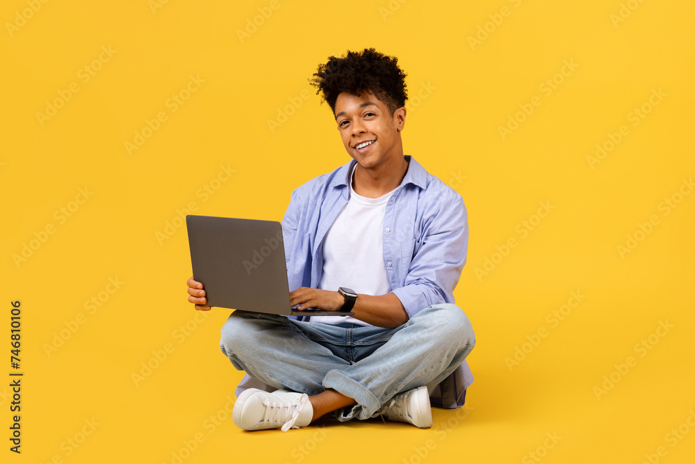 Smiling black student guy with laptop sitting cross-legged on yellow background Smiling black student guy with laptop sitting cross-legged on yellow background