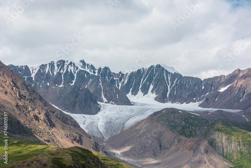 Fototapeta Naklejka Na Ścianę i Meble -  Dramatic landscape with glacier tongue among rocky ridges and sharp snow-capped mountain range under gray cloudy sky. Alpine valley among green hills and rocks in sunlight against ice and sheer crags.
