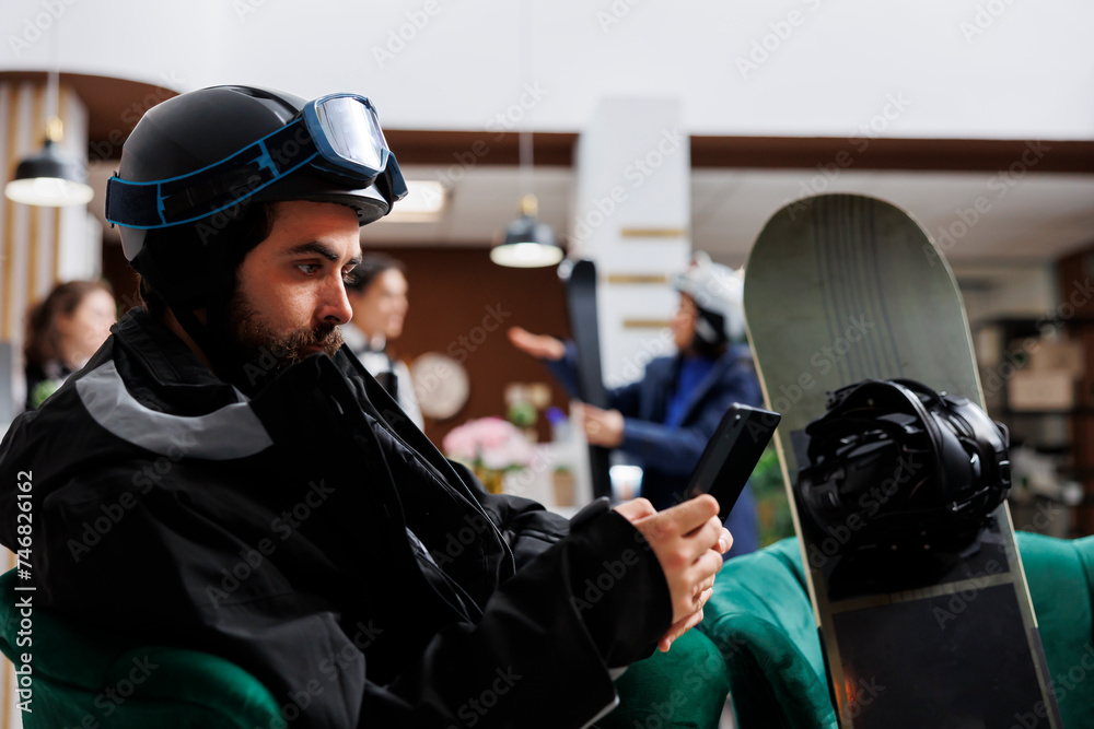 Male tourist in winter attire relaxes on sofa in hotel lobby, browsing on digital tablet. Caucasian man with snowboarding gear makes reservation with smart device for winter holiday at ski resort. Male tourist in winter attire relaxes on sofa in hotel lobby, browsing on digital tablet. Caucasian man with snowboarding gear makes reservation with smart device for winter holiday at ski resort.