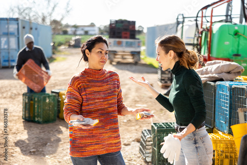 Emotional displeased young female farmer reprimanding confused apologetic asian workwoman for unfulfilled job at farm on sunny spring day photo