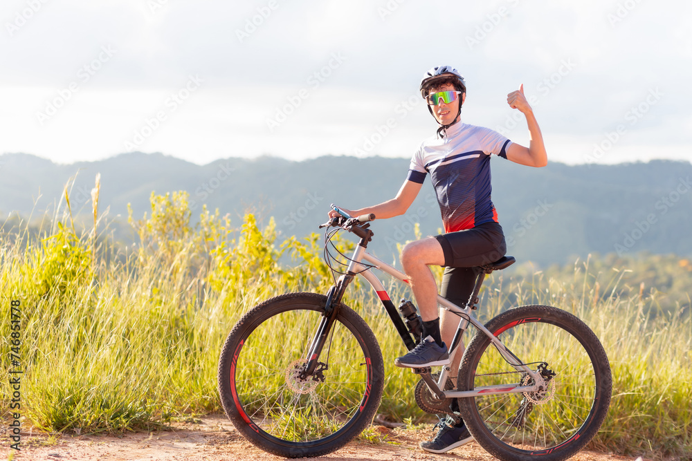 Fototapeta premium Brazilian cyclist standing with his bike and giving a thumbs up sign with his hand. He is happy and smiling while looking at the camera. In the background, landscapes with hills.
