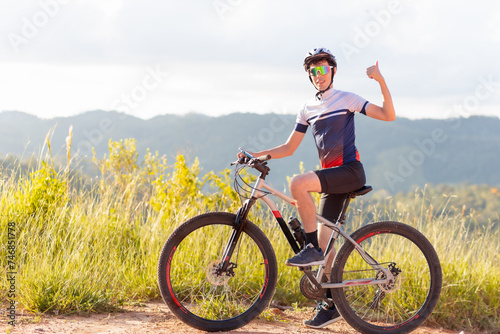 Brazilian cyclist standing with his bike and giving a thumbs up sign with his hand. He is happy and smiling while looking at the camera. In the background, landscapes with hills.