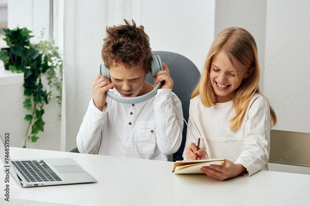 Happy Caucasian boy and girl studying at home together, sitting at a table in the living room They are using laptops for online elearning, wearing headphones and typing on their keyboards The room is