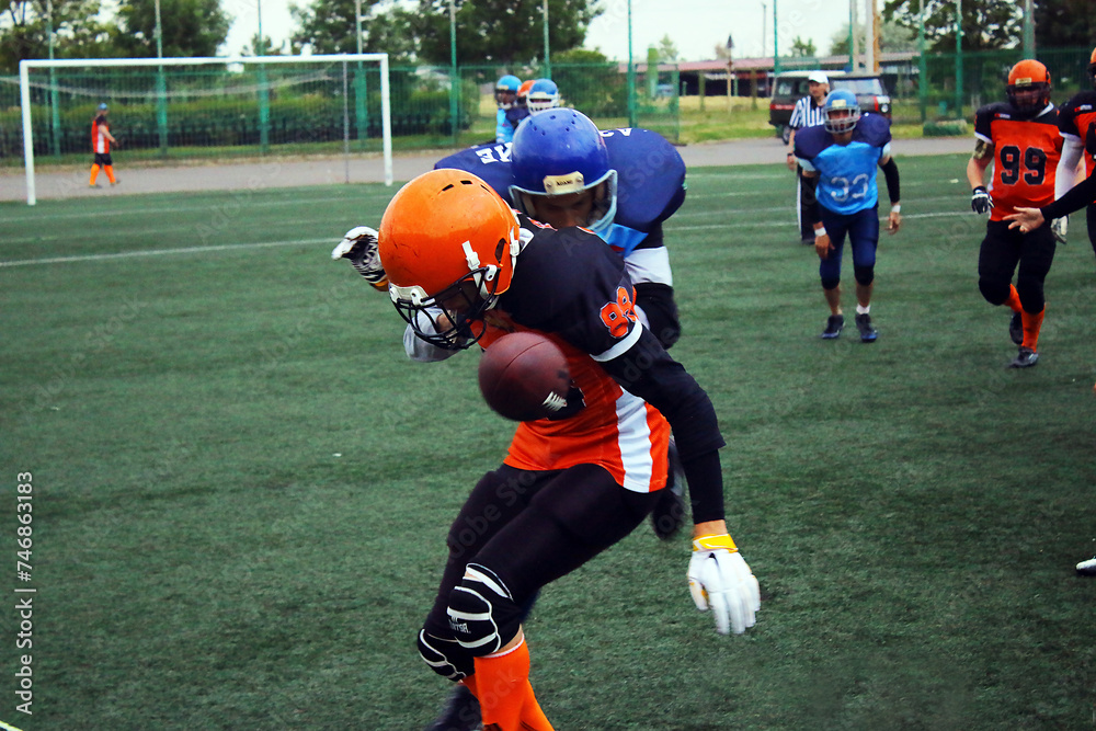 An American football player in a black and blue uniform knocks down a player in a black and orange uniform on a green field