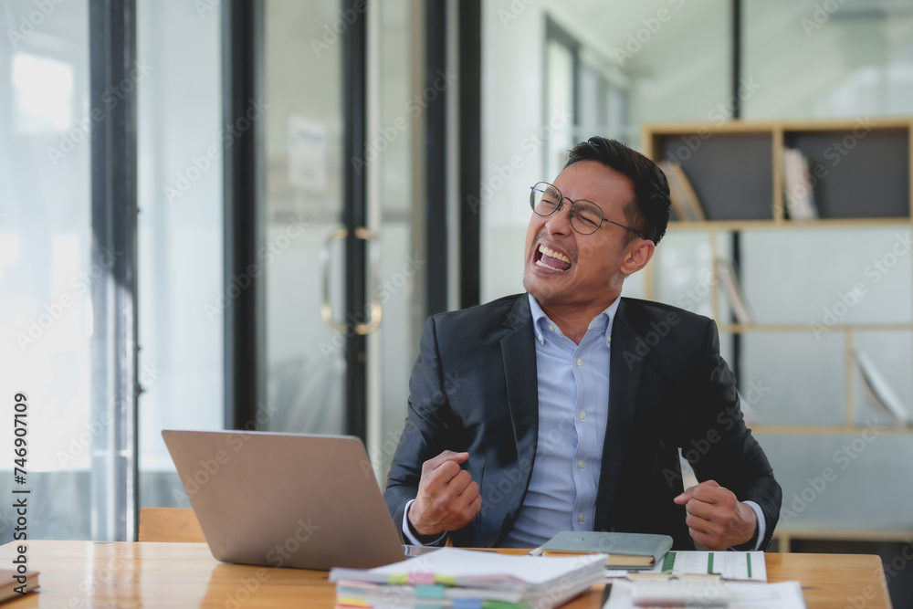 Happy asian businessman celebrating success with arms raised with laptop computer at office desk. Achievement and job satisfaction concept.