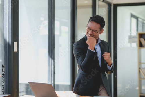 Happy asian businessman celebrating success with arms raised with laptop computer at office desk. Achievement and job satisfaction concept.