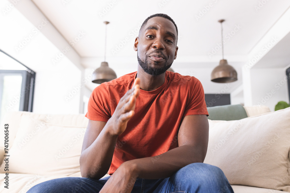 African American man in a red shirt is sitting on a couch, gesturing with his hand on a video call