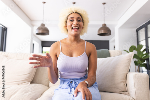 Young biracial woman with curly blonde hair smiles and gestures while sitting on couch on video call