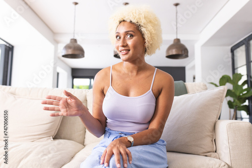 Young biracial woman with curly blonde hair sits on couch, gesturing mid-conversation on video call