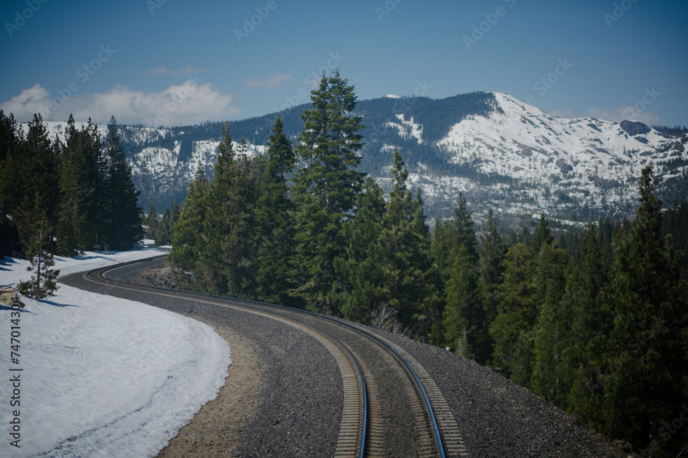 Traveling along a railway in the Sierra Nevada mountains near Truckee, California, a view  of snow-covered mountains, a pine forest, and the train tracks themselves curving through the scenery.