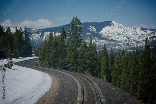 Traveling along a railway in the Sierra Nevada mountains near Truckee, California, a view of snow-covered mountains, a pine forest, and the train tracks themselves curving through the scenery.