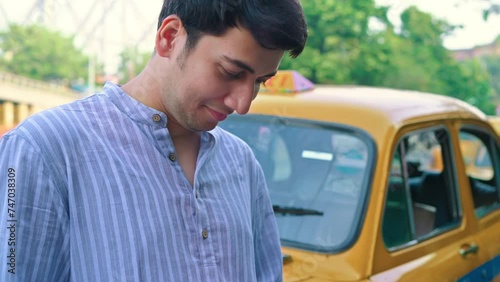 A young smiling Indian Asian good looking man or male is resting on an iconic or vintage yellow cab outdoors and using a mobile phone or smartphone with Howrah Bridge, Kolkata in the background