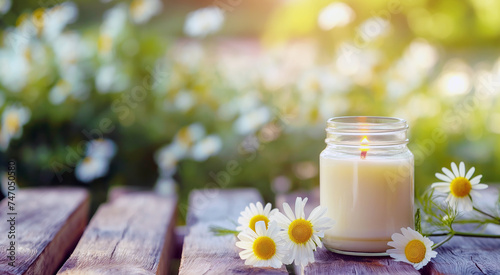 Candle in jar with lit flame on wooden surface among fresh daisies in soft-focused floral background