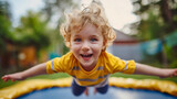 Closeup of the happy toddler boy with curly blonde hair, flying in the air, jumping on the trampoline outdoors in his backyard, childhood leisure time activities on a sunny summer day outdoors