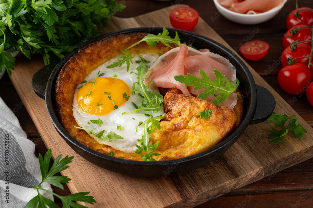 Savory Dutch baby pancake with cheese, fried egg, arugula and prosciutto in cast iron pan on a dark wooden background. Savory Dutch baby pancake with cheese, fried egg, arugula and prosciutto in cast iron pan on a dark wooden background.