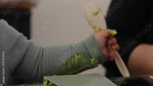 Baby boy with messy clothes holds a spoonful of avocado