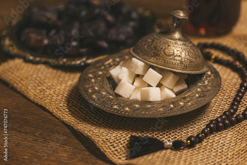 Sugar cubes in a bronze plate, dried dates and tea on a wooden background with burlap. Ramadan Kareem holiday background. Islamic Holy Month Greeting Card. Soft focus. Shallow DOF.