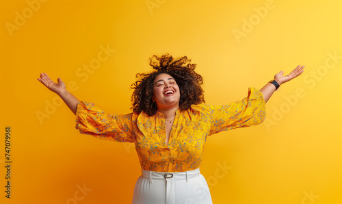 Curvy woman in joyful movement, smiling with arms raised in a positive pose against a vibrant yellow background, radiating energy and happiness.