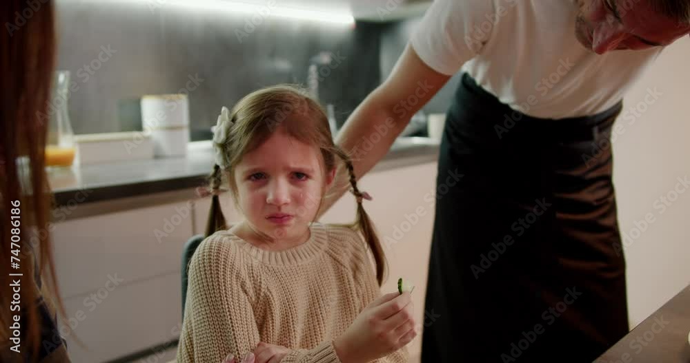Sad little girl with tears in her eyes talks about her experiences and difficulties to her mom and dad during dinner near the family table in the kitchen in the evening Sad little girl with tears in her eyes talks about her experiences and difficulties to her mom and dad during dinner near the family table in the kitchen in the evening