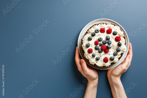 Woman hands holding sponge cake decorated whipped cream, raspberries and blueberries on a blue background. Copy space. Top view. photo