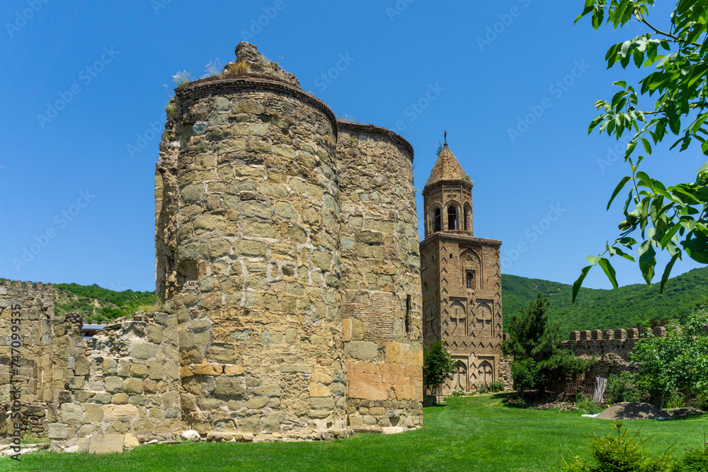 Monastery courtyard. Ruins of old church on the green grass. Stone wall of tower. Brick bell tower. Daylight, blue sky.