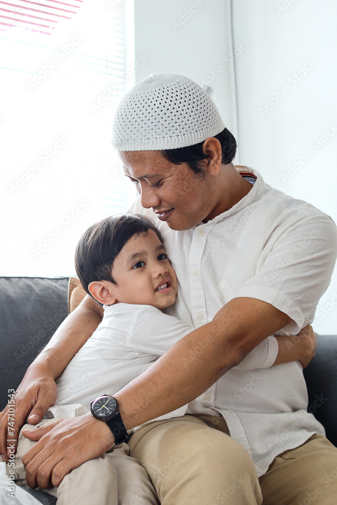 Muslim child boy hugging father during Eid Mubarak while sitting together on the sofa Muslim child boy hugging father during Eid Mubarak while sitting together on the sofa