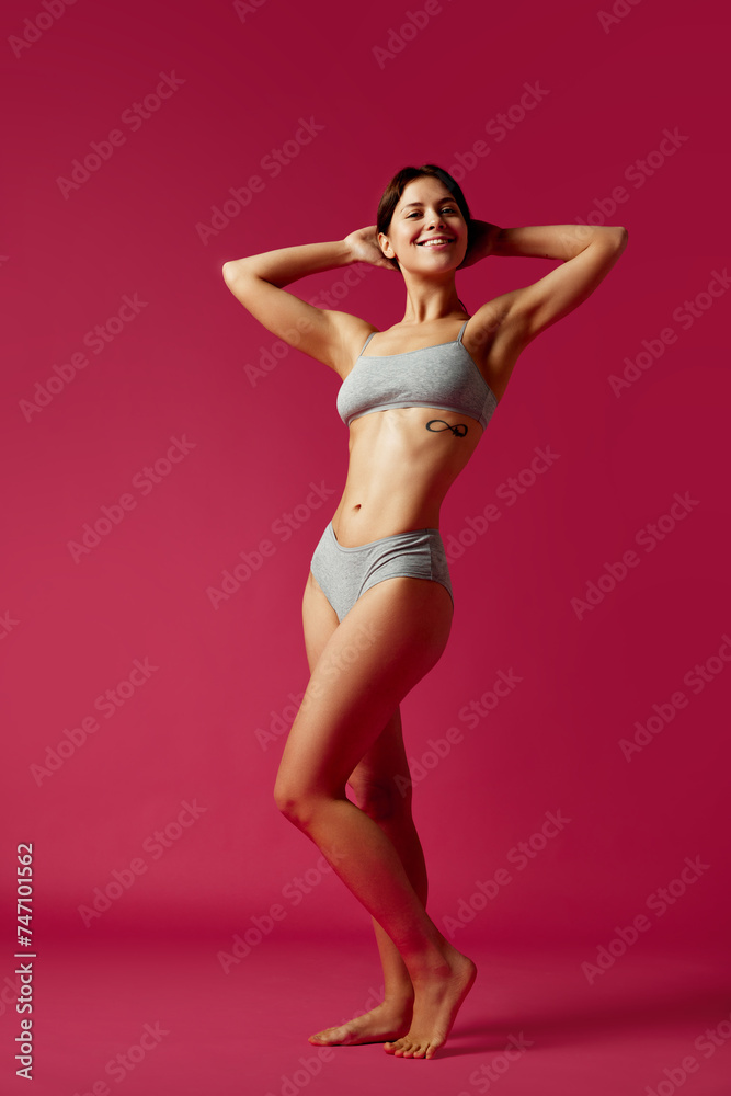 Full-length portrait of young smiling woman posing raising hands demonstrating her perfect body curves against vivid pink background. Concept of beauty treatments, dieting, female health, femininity.