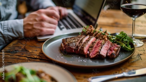 
A person is sitting at a table working on a laptop. On the table next to the laptop is a plate with a steak cut into slices on it