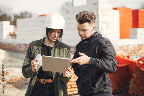 Stylish woman architect with tablet and contractor man checking blueprints at construction site. Young engineer or construction workers in hardhat looking at plans of new modern house