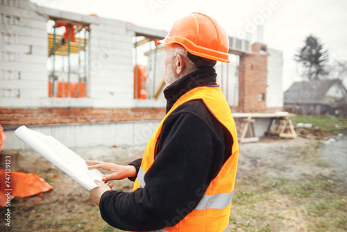 Senior male foreman in hardhat checking blueprints of building new modern house. Man engineer or construction worker looking at plans at construction site. Copy space