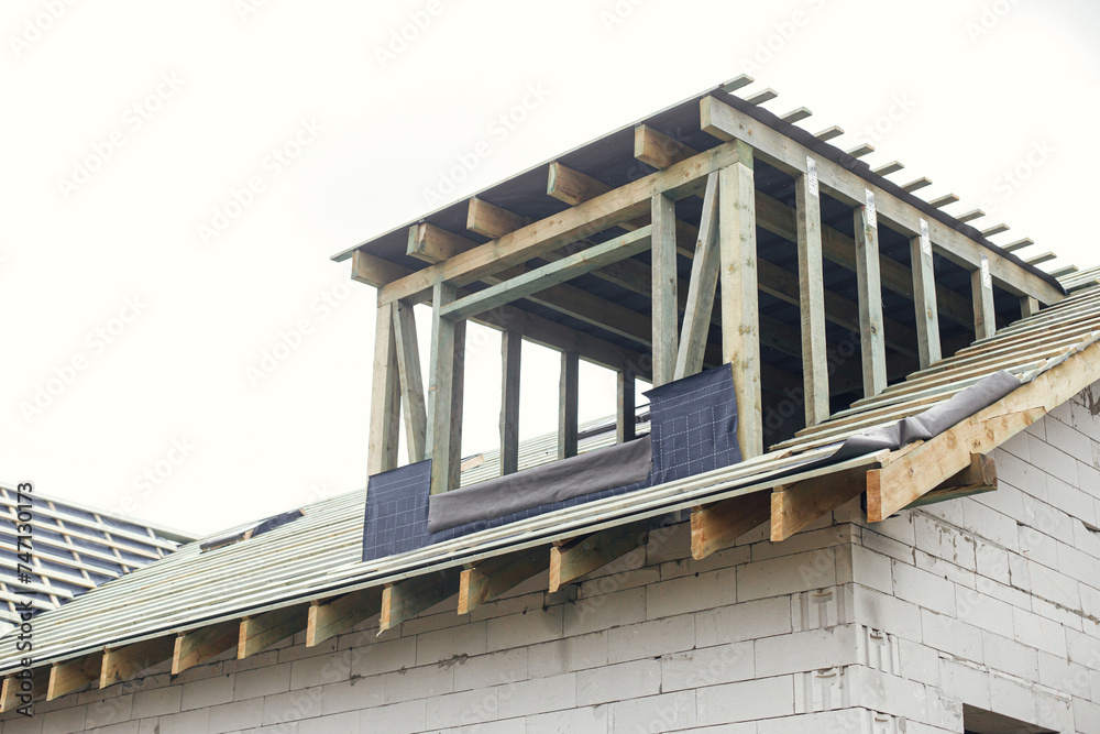 Unfinished house. Wooden roof framing with vapor barrier and dormer on block walls with windows. New modern farmhouse construction. Timber trusses Rafters and beams on aerated concrete blocks