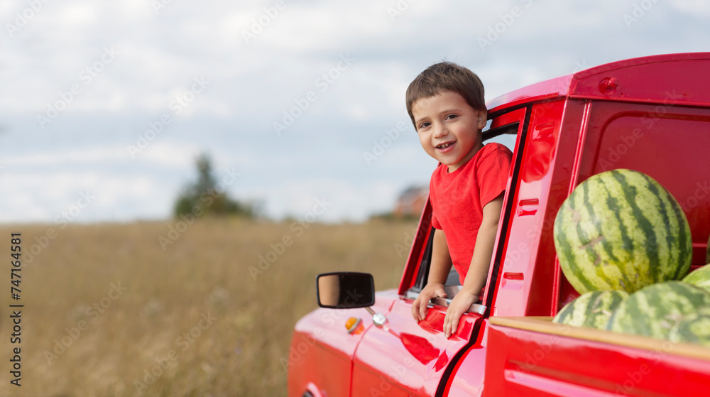 child boy waves from the cab of a red pickup truck with watermelons. banner. copy space.summer time in nature in the village child boy waves from the cab of a red pickup truck with watermelons. banner. copy space.summer time in nature in the village