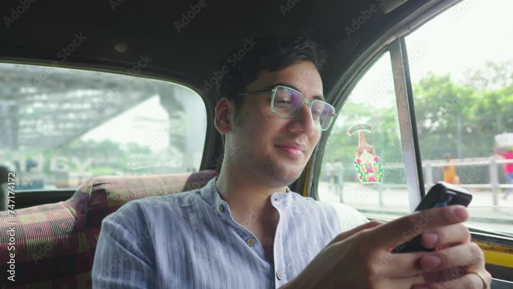 A young attractive smiling Indian Asian man or male sitting in a moving car or cab using a mobile phone or smartphone to type a text message with the iconic Howrah bridge is visible in the background