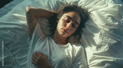 A woman with her eyes closed appears to be in a state of deep relaxation or sleep lying on her back with her arms crossed over her chest on a bed with white sheets and pillows. photo