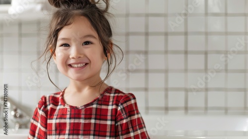 Smiling young girl with dark hair wearing red and white plaid shirt standing in front of white tiled wall with silver faucet.