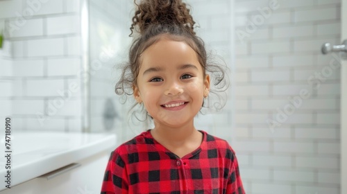 Smiling young girl with curly hair wearing red and black checkered pajamas standing in a white tiled bathroom with a sink in the background.