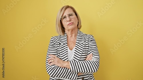Disgruntled middle-age blonde woman in striped jacket stands crossed-armed, expressing nervous skepticism and disapproval. negatively charged personality isolated on vibrant yellow background.