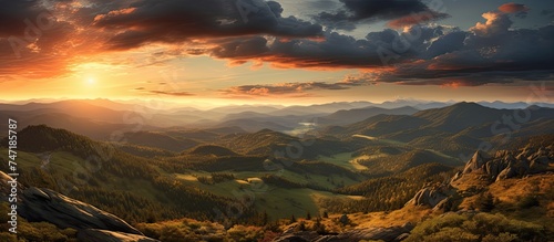 The painting depicts a vibrant sunset over the Czech Republics Orlicke hory mountain range. The sky is ablaze with hues of orange, pink, and purple, casting a warm glow over the rugged peaks. photo