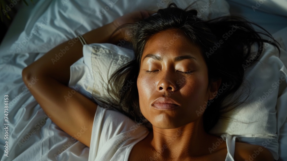 A woman with dark hair lying on her back with her eyes closed wearing a white top resting on a white bed with white sheets and pillows. A woman with dark hair lying on her back with her eyes closed wearing a white top resting on a white bed with white sheets and pillows.