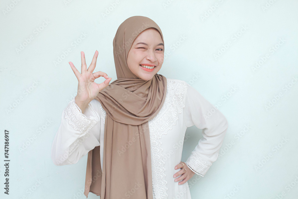 Young Asian Muslim woman wearing hijab, smiling and see camera with ok gesture. Isolated in gray background. Young Asian Muslim woman wearing hijab, smiling and see camera with ok gesture. Isolated in gray background.
