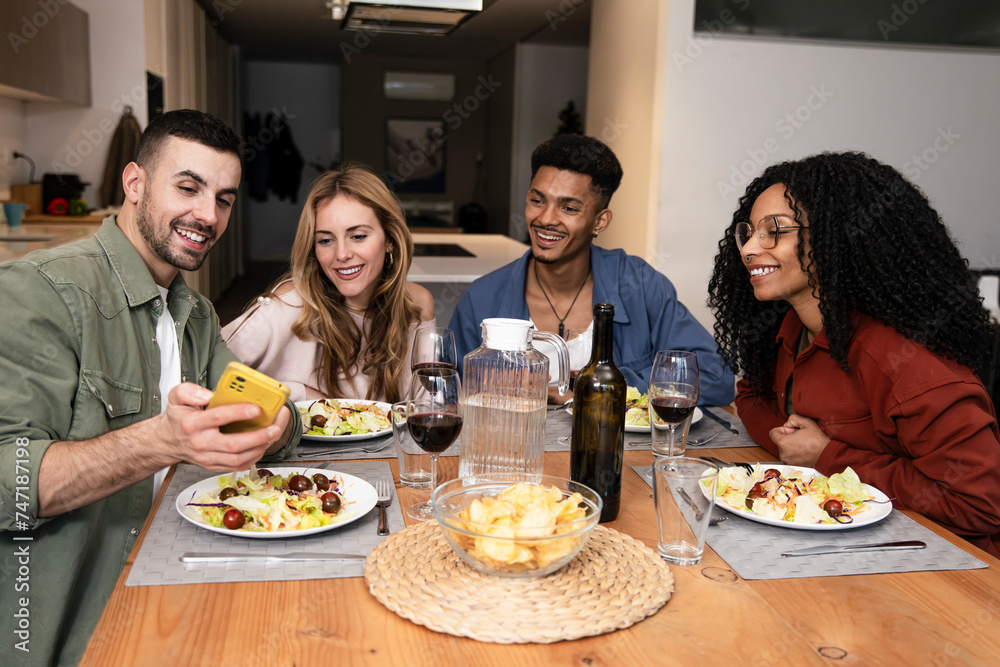 Smiling people sharing smartphone while eating on a modern apartment. Diverse group of happy friends having video call diner at home. 