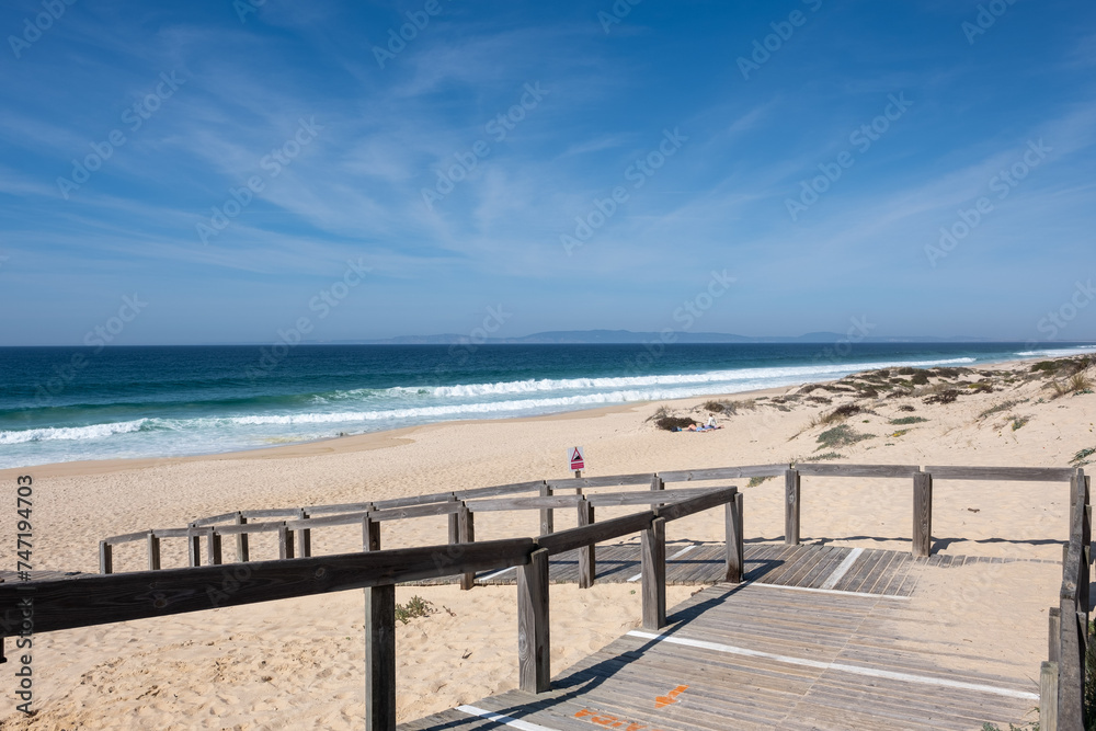 A tranquil beach scene in Comporta Beach, Setubal, Portugal, with a wooden boardwalk leading to a sandy beach against a backdrop of a clear blue sky and rolling waves