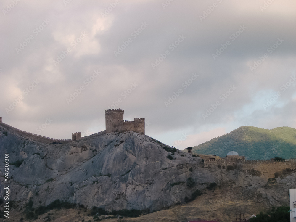 An ancient medieval fortress on the edge of a cliff is surrounded by large, bright clouds. Landscape of a tourist and historical place with a cold blue tint.